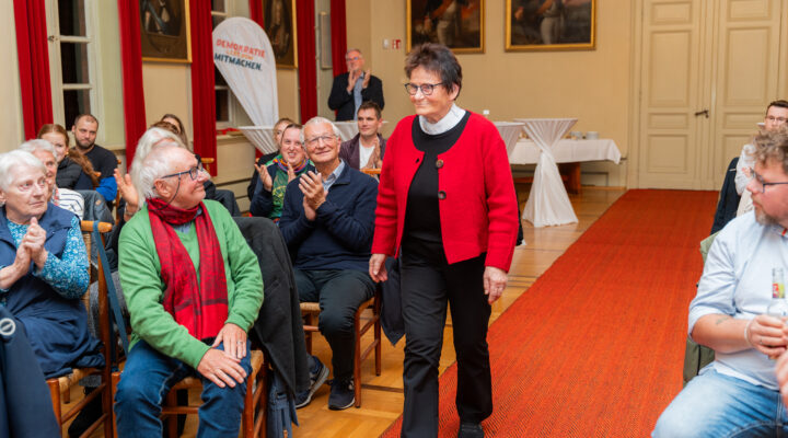 Rosi Knocke, Trägerin eines Sonderpreises beim Carl-Osterwald-Preis 2026, steht lächelnd im Veranstaltungssaal. Die um sie herum sitzenden Gäste applaudieren ihr.
