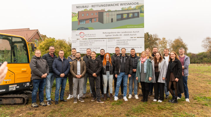 Kolleginnen und Kollegen des Rettungsdienstes des Landkreises Aurich stehen gemeinsam vor einer großen Bautafel mit einer Visualisierung der geplanten neuen Rettungswache in Wiesmoor. Links ist ein gelber Bagger zu sehen.