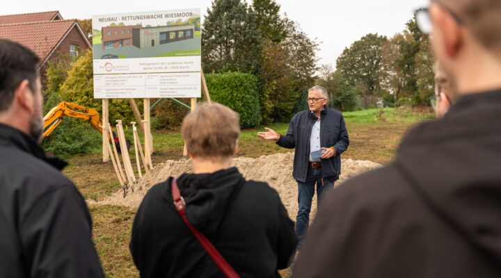 Landrat Olaf Meinen spricht beim offiziellen Spatenstich für den Neubau der Rettungswache Wiesmoor zu den anwesenden Gästen. Im Hintergrund sind eine Bautafel mit der Visualisierung des geplanten Gebäudes, mehrere Spaten und ein gelber Bagger zu sehen.
