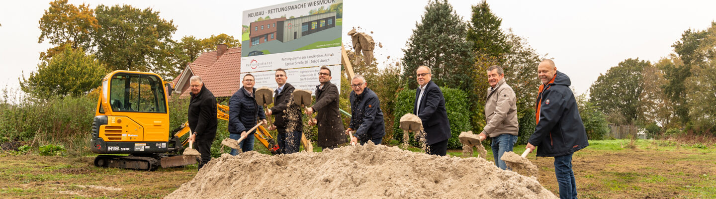 Acht Männer führen mit Spaten den symbolischen ersten Spatenstich für den Neubau der Rettungswache Wiesmoor durch. Im Hintergrund stehen eine Bautafel mit einer Visualisierung des geplanten Gebäudes und ein gelber Bagger.
