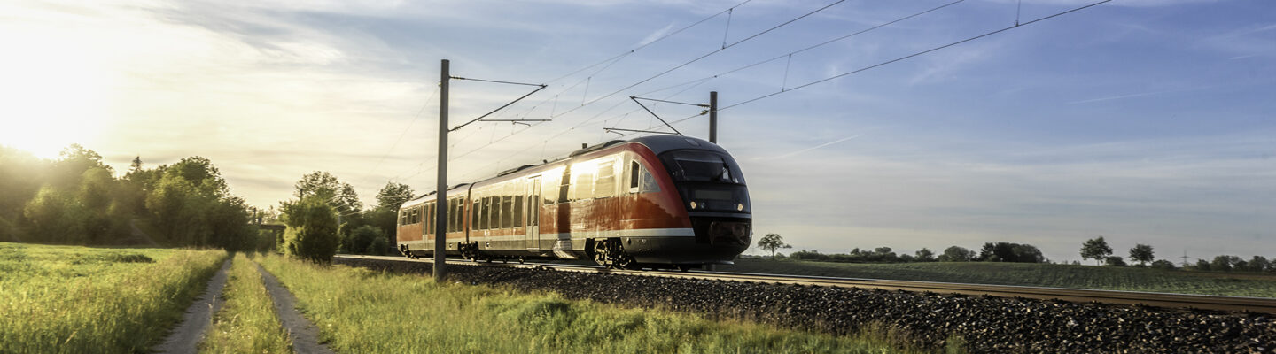 Ein rot-weißer Regionalzug fährt auf einer elektrifizierten Bahnstrecke durch eine ländliche Landschaft. Neben den Gleisen verläuft ein schmaler Feldweg durch grüne Wiesen. Die tief stehende Sonne taucht die Landschaft unter einem leicht bewölkten, blauen Himmel in warmes Licht.