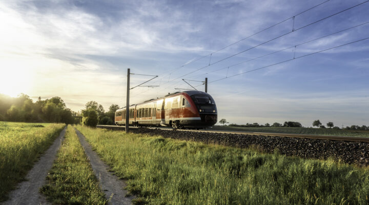 Ein rot-weißer Regionalzug fährt auf einer elektrifizierten Bahnstrecke durch eine ländliche Landschaft. Neben den Gleisen verläuft ein schmaler Feldweg durch grüne Wiesen. Die tief stehende Sonne taucht die Landschaft unter einem leicht bewölkten, blauen Himmel in warmes Licht.